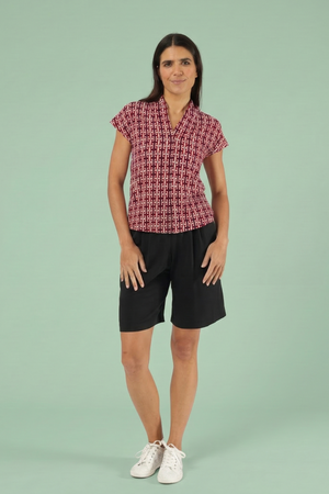 Frau trägt rote Kurzarmbluse mit geometrischem Muster, kombiniert mit schwarzen Shorts und weißen Sneakern vor einem hellgrünen Hintergrund.