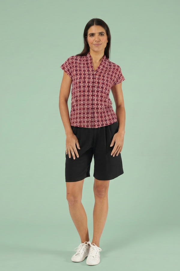 Frau trägt rote Kurzarmbluse mit geometrischem Muster, kombiniert mit schwarzen Shorts und weißen Sneakern vor einem hellgrünen Hintergrund.